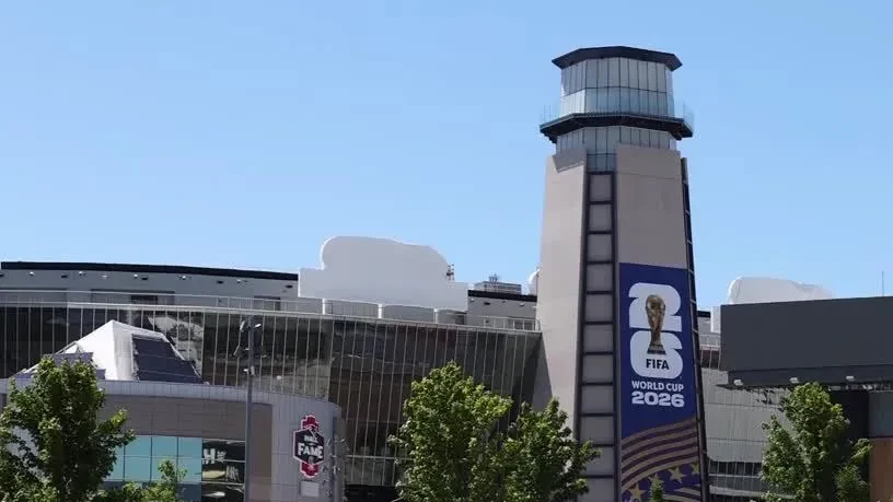 Torcida em estádio durante dia de calor, com bandeiras e torcedores se abrigando do sol