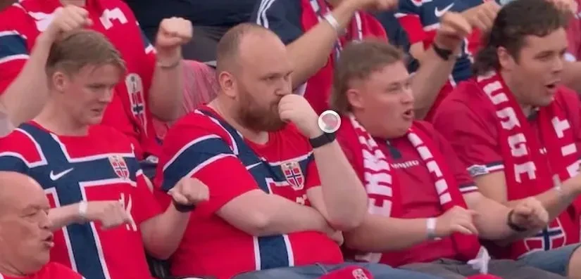 Torcedor norueguês sentado no estádio cercado por torcedores fazendo a remada viking