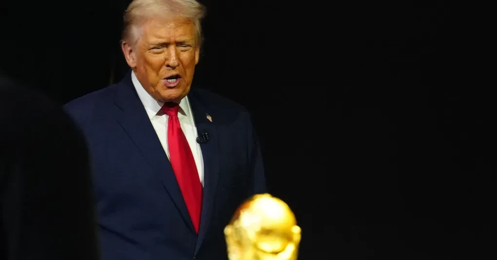 Donald Trump em evento oficial, acenando com bandeira dos Estados Unidos ao fundo