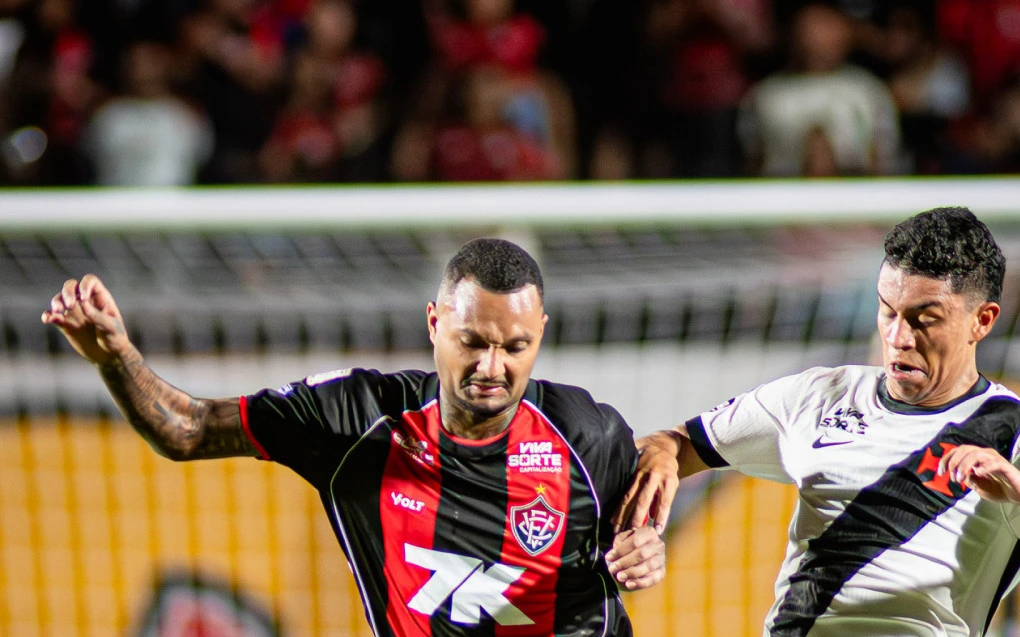 Jogadores disputam bola no Barradão durante jogo entre Vitória e Vasco, com torcida ao fundo